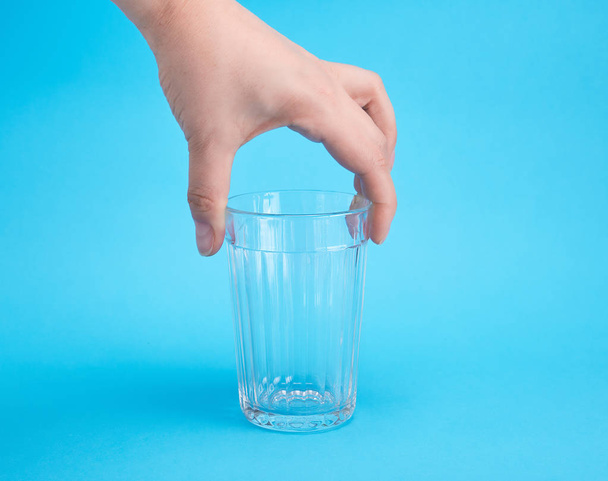 Die Hand der Frau hält ein leeres transparentes Glas, blauer Hintergrund - Foto, Bild