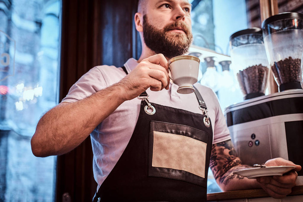 Barista guapo en delantal bebiendo café durante la pausa para el almuerzo apoyado en un mostrador en la cafetería
 - Foto, imagen