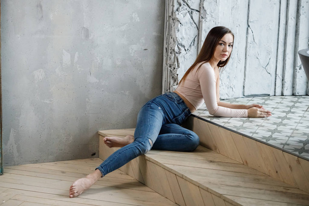 Портрет красивої молодої жінки в блакитних джинсах і бежевій футболці. Молода модель сидить на підлозі в інтер'єрі розкішних квартир
 - Фото, зображення
