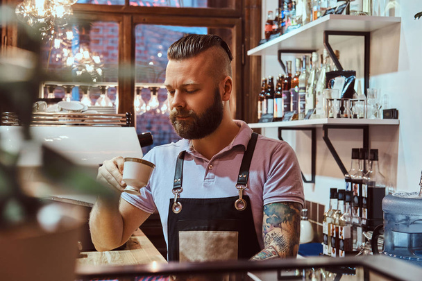 Бариста в фартуке пьет кофе во время обеденного перерыва, стоя за стойкой в кафе
 - Фото, изображение