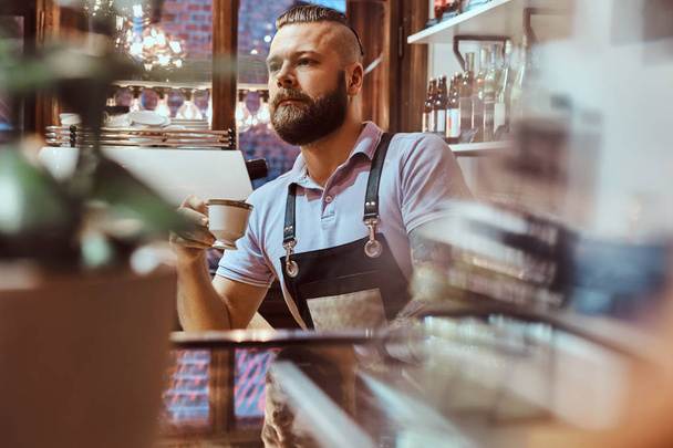 Barista στην ποδιά πίνοντας καφέ κατά τη διάρκεια του μεσημεριανού διαλείμματος που στέκεται πίσω από τον πάγκο στο κατάστημα καφέ - Φωτογραφία, εικόνα