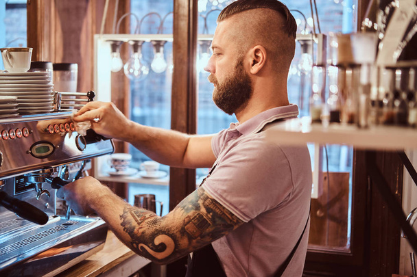 tätowierter Barista mit stylischem Bart und Frisur bei der Arbeit an einer Kaffeemaschine in einem Café oder Restaurant - Foto, Bild