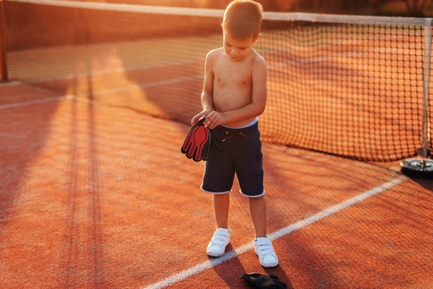 Shirtless sporty boy putting on goalkeeper's gloves while standing on the playground in the morning at summertime. - Photo, Image