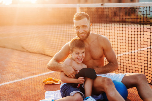 Hymyilevä paidaton isä halaa poikaansa istuessaan kentällä kesällä.
. - Valokuva, kuva