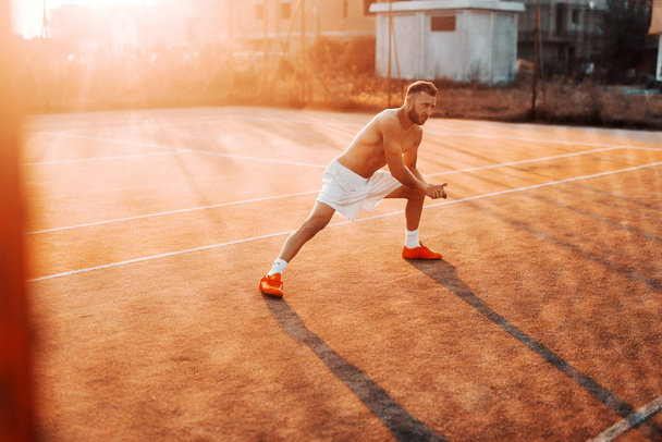 Shirtless bearded Caucasian man in shorts stretching leg before exercising on the court in the morning at summer. - Fotoğraf, Görsel