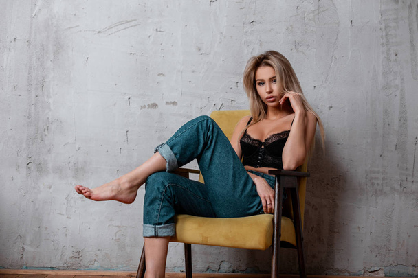 Charming attractive young woman with blond long hair with natural make-up in a luxurious black lace sconce in fashionable blue jeans poses on a vintage chair while sitting in a room. Elegant sexy girl - Φωτογραφία, εικόνα
