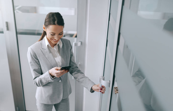 Jolie brune caucasienne en tenue formelle utilisant un téléphone intelligent et entrant dans le bureau. Chaque jour est une chance d'être meilleur
. - Photo, image