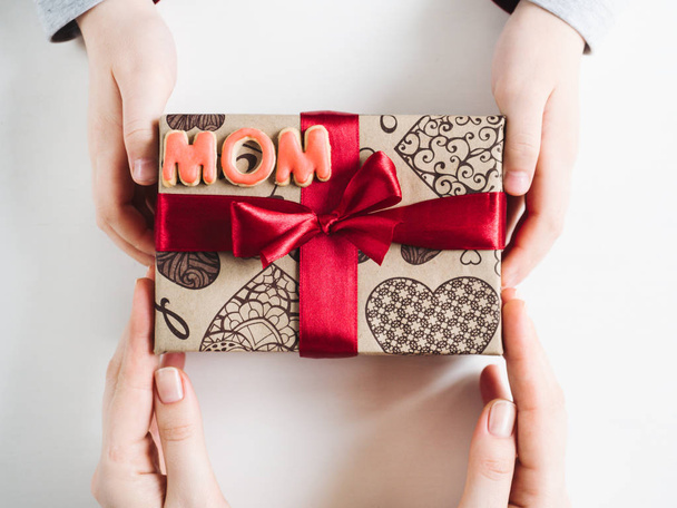 Children's hands, beautiful box with a gift, red ribbon and the word MOM on a white, isolated background. Top view, close-up. Preparing for the holidays - Foto, afbeelding