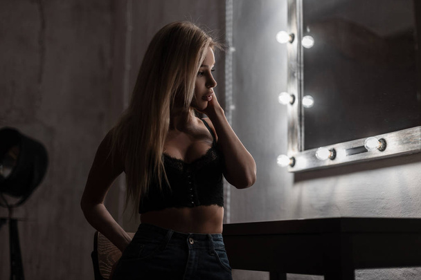 Sexy charming young woman with beautiful body with blond hair in a stylish lace bra in fashionable jeans is standing in a dark bedroom near a vintage wooden mirror with light bulbs. Pretty girl. - Photo, Image
