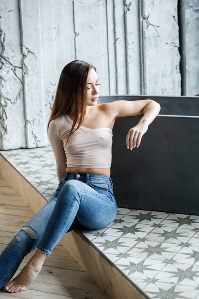 Portrait of beautiful young woman in blue jeans and beige t-shirt. Young model sitting on floor at luxury apartments interior - Photo, Image