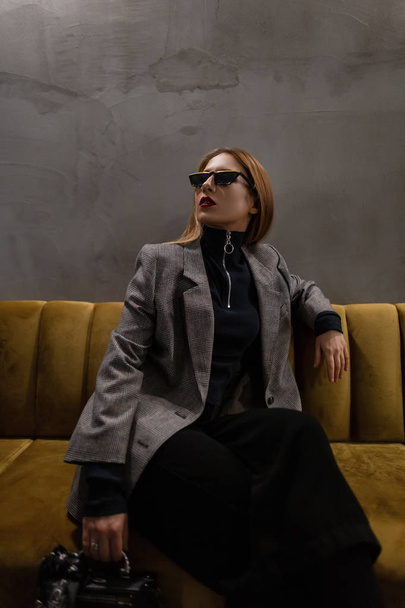 Stylish hipster young woman in sunglasses with a pierced nose with red lips in an elegant checkered jacket in black pants with a vintage handbag-recorder sitting on a sofa in the room. Cool girl. - Foto, Bild