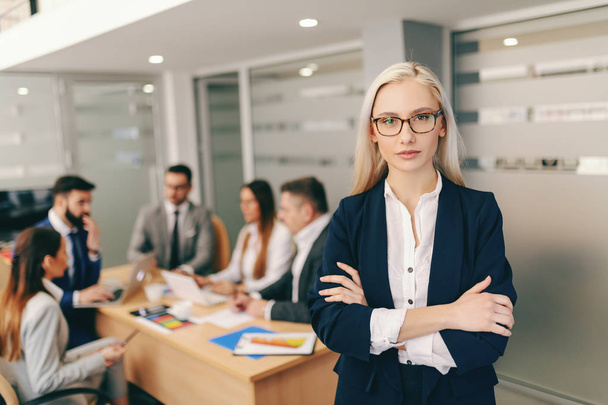 Portrait of attractive blonde businesswoman in formal wear holding crossed arms while standing in board room In background colleagues having meeting - Foto, Imagen