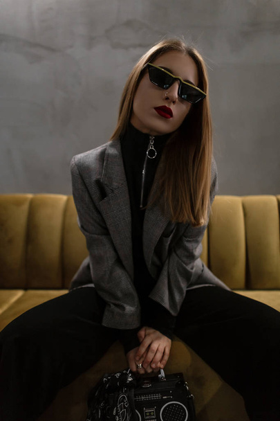 Mooie moderne jonge vrouwen hipster met doordrongen neus met rode lippen in een vintage grijs geruit jasje in zwarte broek met een handtas-recorder zittend op een bank in een café. Stijlvolle ouderwetse meisje. - Foto, afbeelding