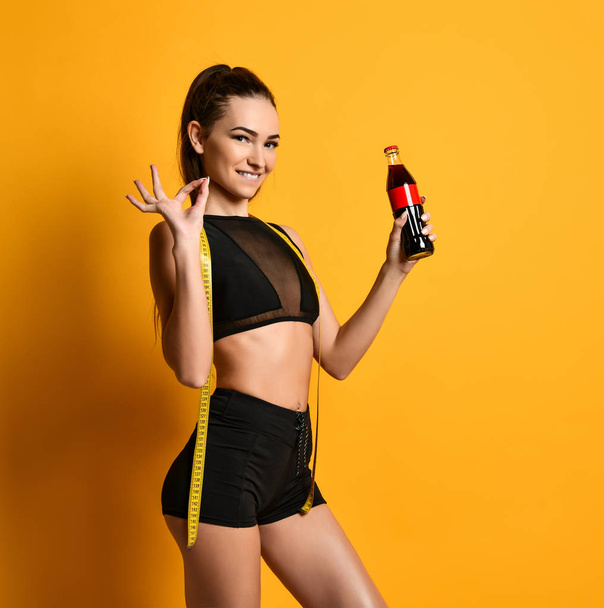 nice young fitness girl in black sportswear is looking at the camera and keeps at hand a tape measure and a bottle of sports nutrition liquid. Healthy life weight loss concept - Φωτογραφία, εικόνα