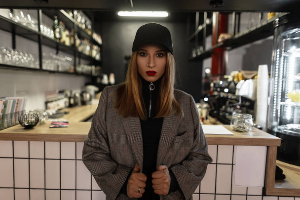 Vonzó fiatal stílusos nő divatos kap egy retro stílusú vintage kabát kockás piros ajkak orr piercing áll egy kávézóban, a háttérben a bár a számláló. Jó régimódi lány. - Fotó, kép