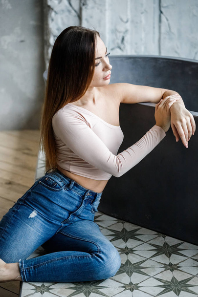 Portrait of beautiful young woman in blue jeans and beige t-shirt. Young model sitting on floor at luxury apartments interior - Foto, Bild