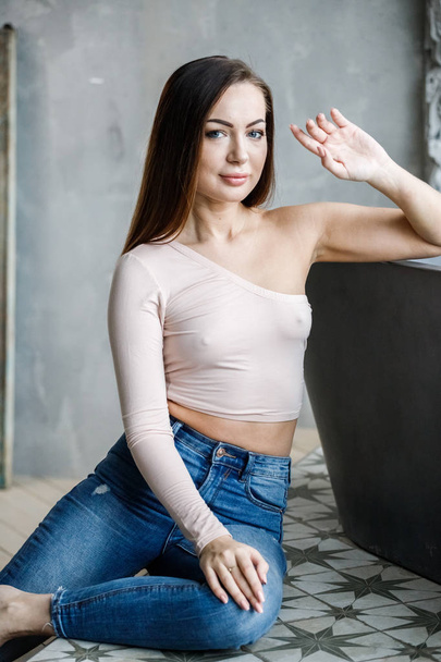 Portrait of beautiful young woman in blue jeans and beige t-shirt. Young model sitting on floor at luxury apartments interior - Fotografie, Obrázek