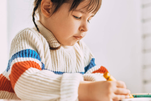 Closeup image portrait of creative cute little girl playing with oil pencils, sitting at white desk at home. Pretty preschool kid draws with colorful pencils. People, childhood and education concept - Fotó, kép