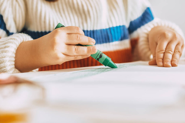 Cropped image of cute little girl paints with green oil pencil sitting at white desk at home. Image of pretty preschool kid draws on paper in the kindergarten. People, childhood and education concept - Foto, Bild
