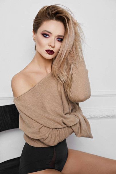 Portrait of sexy and beautiful blonde model girl with bright makeup and amazing blue eyes in black stylish lingerie and in sweatshirt sits on a chair and posing at pink background - Φωτογραφία, εικόνα