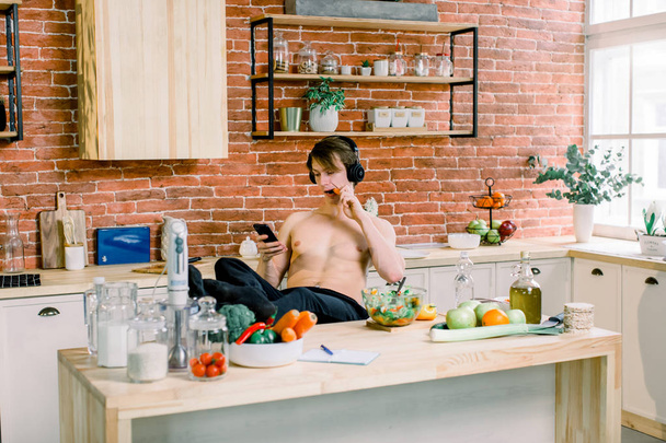 Handsome man cooking at home preparing salad in kitchen. Young man is listening to music in earphones, using smartphone and eating carrot while preparing salad. Healthy lifestyle and food - Photo, image