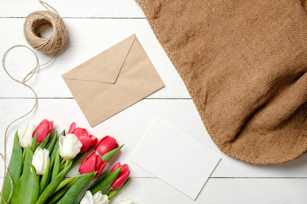 Bouquet of tulips flowers, kraft envelope, twine, burlap on white wooden table. Vintage greeting card for womans day, mothers day, birthday, easter. Rustic background, flat lay, above view. - 写真・画像