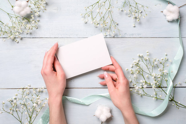 Hands holding blank paper card on light blue wooden desk with flowers. Tender greeting card for womens or mothers day, easter, spring holidays. - Фото, зображення