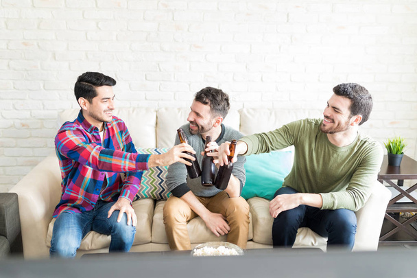 Веселые друзья-мужчины звонят в бутылки пива, сидя дома на диване
 - Фото, изображение