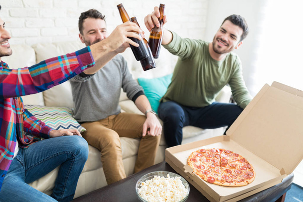 Happy male friends toasting beer bottles with pizza and popcorn on table at home - Foto, afbeelding