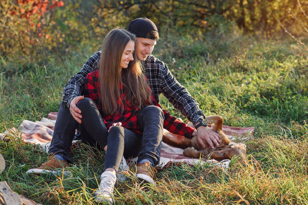 Happy caucasian couple with guitar resting on picnic in spring park. Young man and his girlfriend playing with their funny dog. - Φωτογραφία, εικόνα