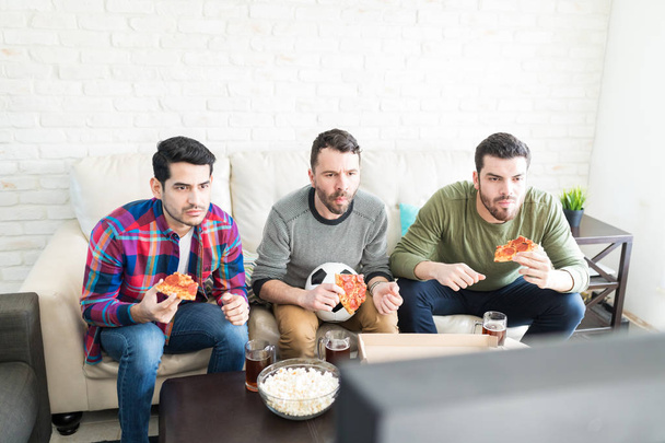 Фокусированные футбольные болельщики смотрят телевизор и едят фаст-фуд дома
 - Фото, изображение