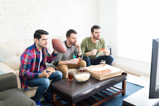 ernsthafte gutaussehende Männer, die ein amerikanisches Fußballspiel im Fernsehen verfolgen, während sie zu Hause Pizza und Bier trinken - Foto, Bild
