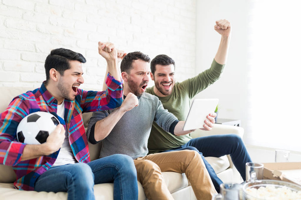 Male friends cheering while watching live streaming of soccer match - Zdjęcie, obraz