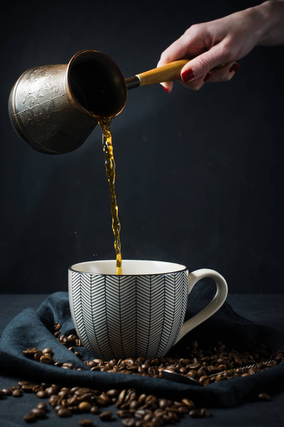 Женская рука наливает кофе из тюрков в чашку. Черный фон, вид сверху, место для текста
 - Фото, изображение
