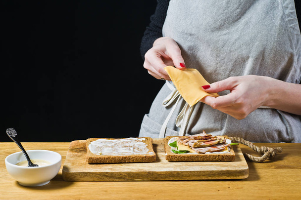 The chef prepares a sandwich, puts slices of cheese on toast. Black background, side view, kitchen - Photo, Image