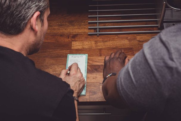 Een Sex tussen verschillendre rassen homo paar maken van een boodschappenlijstje voor de boodschappen boven op een houten keuken thuis.  - Foto, afbeelding