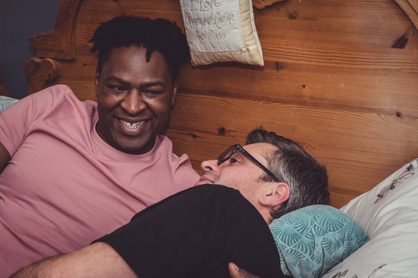 An interracial gay couple talking, lying down in their bedroom.  - Foto, afbeelding
