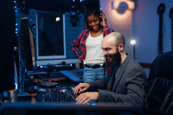 Sound engineer and female singer at remote control panel in audio recording studio. Musician in headphones listens composition, professional music mixing - Photo, Image