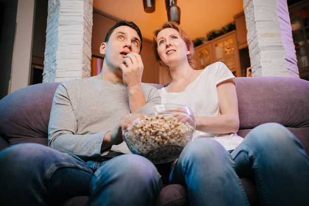 Молодая пара ест попкорн и смотрит кино дома на диване, напуганная
. - Фото, изображение