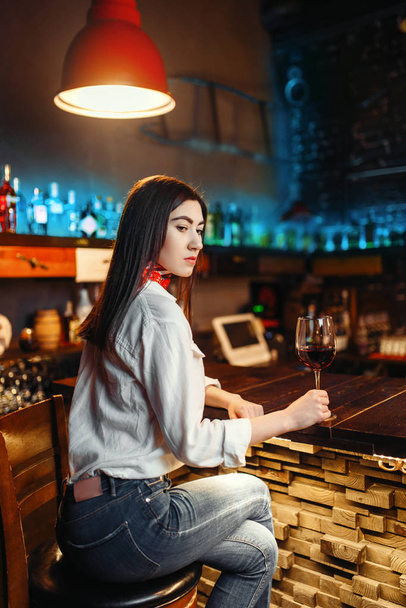 Young lady with glass of red wine sitting at wooden bar counter. Female customer leisures in pub - Foto, immagini