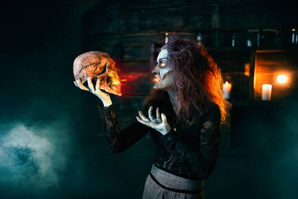 Scary witch holds human skull and reads the spell, dark powers of witchcraft, spiritual seance. Female foreteller calls the spirits, terrible future teller - Photo, Image