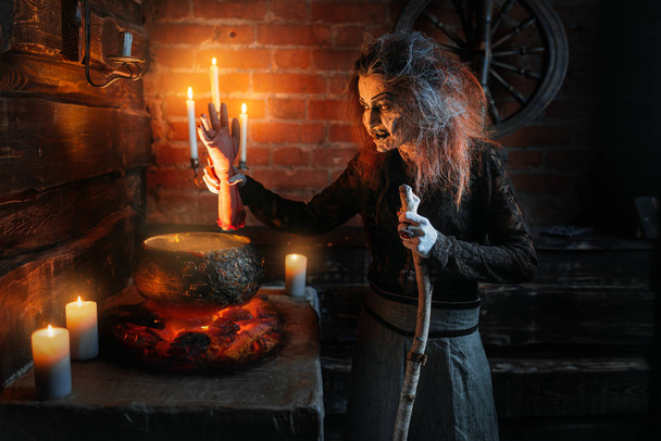 Scary witch reads spell over the pot with human body parts, dark powers of witchcraft, spiritual seance with candles. Female foreteller calls the spirits, terrible future teller - Photo, Image