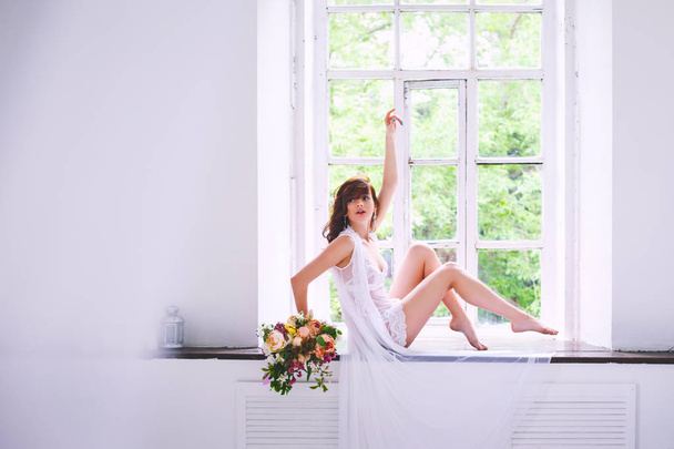 Young bride in a white shirt posing while sitting with a bouquet of flowers on a windowsill - Fotó, kép