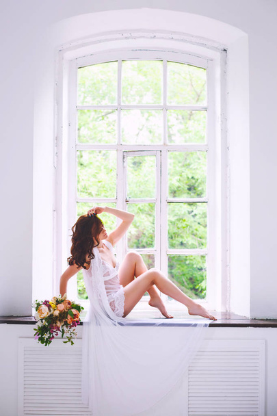 Young bride in a white shirt sits with a bouquet of flowers on a windowsill - Фото, изображение