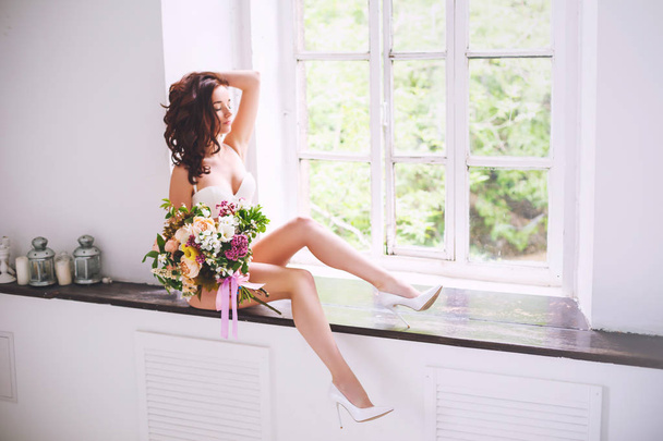 A young bride in a white shirt and a bouquet of flowers looks out the window sitting on the windowsill - Foto, Imagem