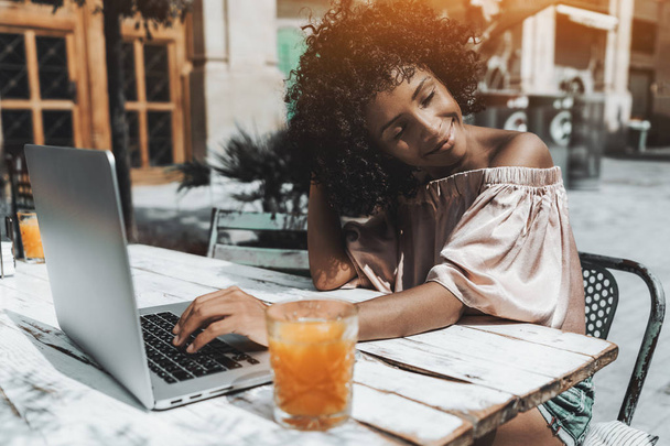 A dazzling young curly hair Brazilian female is enjoying the sun while sitting in a cafe outdoors with her laptop and the glass of orange drink; an African-American girl with netbook in a street bar - Фото, зображення