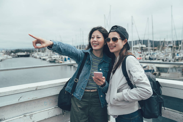 two cheerful sisters travel together sightseeing in Old Fisherman's Wharf in monterey. group of young girl tourist friends sharing beautiful ocean view hand pointing finger far away hold mobile phone - 写真・画像