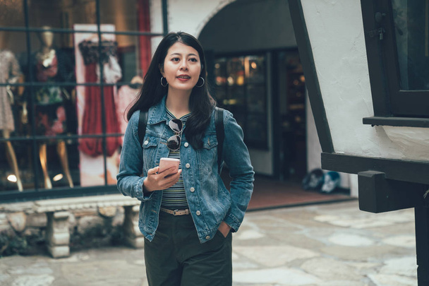 Gyönyörű tizenéves fiatal ázsiai nő a város bevásárló utca-Fashion store ablak. a lány mosolyogva használ smartphone-hoz hálózat online szabadban. női utas gazdaság mobil látogasson el Kármel a tenger - Fotó, kép