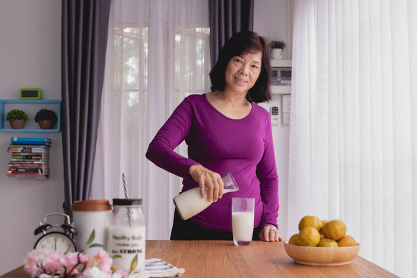アジアの高齢女性が台所のテーブルにミルクを注ぐ。朝食テーブル、正面図. - 写真・画像
