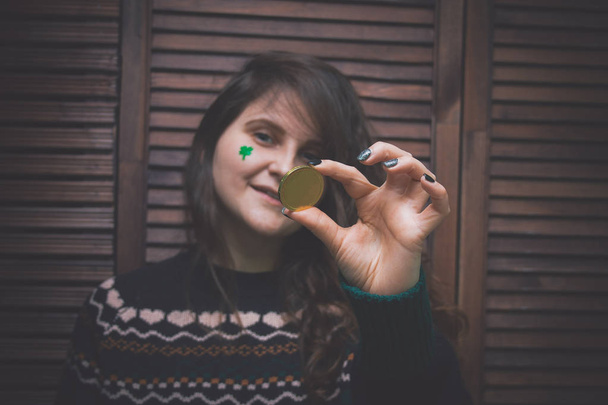 young beautiful woman with painted green clover on her cheek holding golden coin at the bar in front of a wooden wall - Фото, зображення